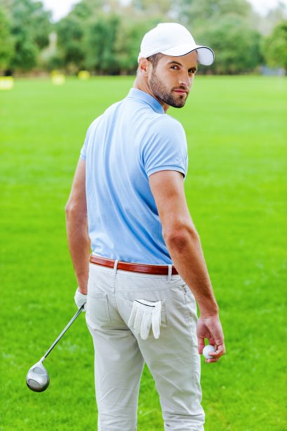 Confident golfer. Rear view of confident golfer holding golf ball and driver while standing on golf course and looking over shoulder