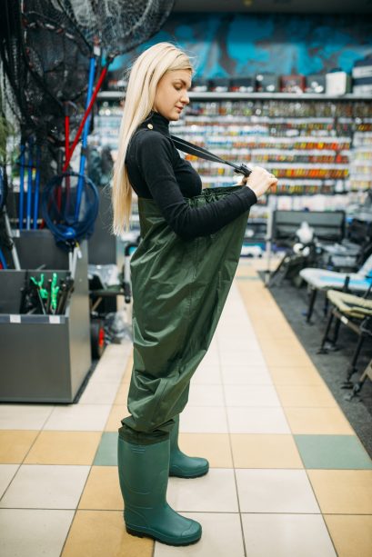 Female angler in large rubber jumpsuit in fishing shop, side view, hooks and baubles on background. Fisherwoman shoosing equipment and tools for fish catching and hunting, accessory in store