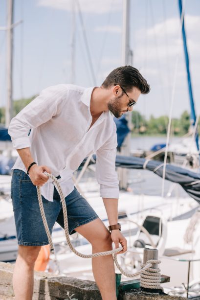 handsome young man in sunglasses holding rope while standing near yacht