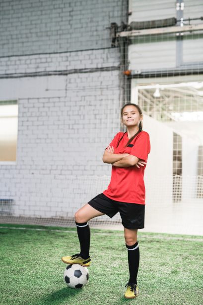 Happy young successful female football player in sports uniform crossing arms by chest and keeping foor on soccer ball