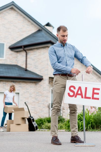 male realtor hanging sale sign in front of woman packing cardboard boxes for relocating from house