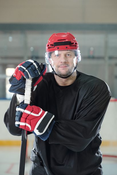 Mature hockey player in sports uniform, protective helmet and gloves standing on rink and looking at you against stadium environment