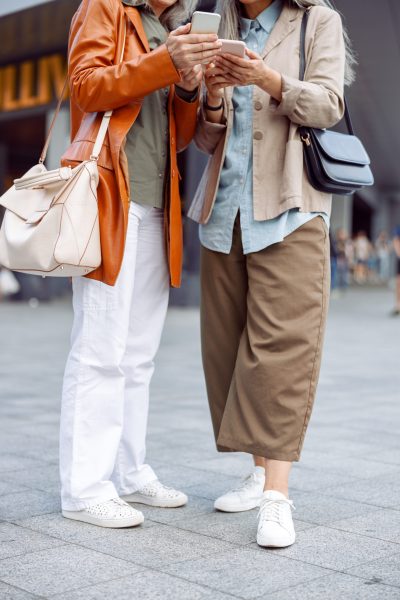 Mature women in stylish apparel hold modern mobile phones standing on city street on autumn day closeup. Old friends meeting