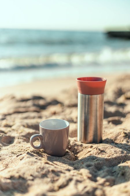 Metal thermos or a thermos mug with coffee or tea drink on the deserted sandy coast of the ocean or sea